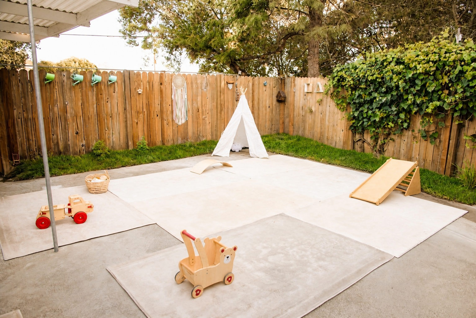 The fenced outdoor play yard with a canvas teepee, wooden ride-on toys, and a climbing ramp