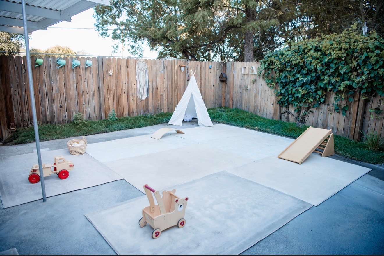 The fenced outdoor play yard with a canvas teepee, wooden ride-on toys, and a climbing ramp